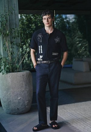 Vīrietis stāv telpās blakus lielam puķu podam ar zaļumiem, valkājot melnu īsroku kreklu ar ziedu rakstu, tumšas bikses, austu jostu un sandales.