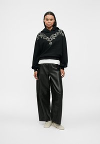 Hanorac negru cu design brodat reprezentând o frunză, asortat cu pantaloni de piele negri supradimensionați și sneakers de culoare deschisă. Cămașă albă simplă vizibilă.