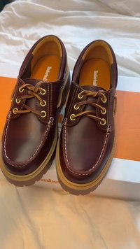 Skórzane buty w ciemnym bordowym kolorze, z złotymi oczkami, kontrastującym przeszyciem i fakturowaną brązową gumową podeszwą. Prezentowane na pomarańczowym pudełku.