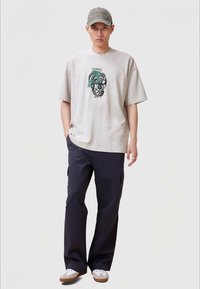 Ανοιχτό γκρι oversized μπλούζα με γραφικό σχέδιο, συνδυασμένη με σκούρες cargo παντελόνες. Το μοντέλο φοράει ένα ταιριαστό γκρι καπέλο και λευκά αθλητικά παπούτσια.