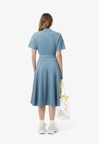 Abito azzurro chiaro con maniche corte, vita cinchata e gonna svasata. Indossato con scarpe da ginnastica bianche e una borsa a tracolla trasparente con rifiniture gialle.