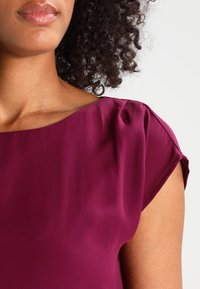 Frau mit lockigem Haar, die eine kurzärmlige, tief magentafarbene Bluse trägt, die Nacken, Schulter und einen Teil ihrer Lippen zeigt.