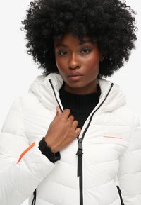 Veste coupe-vent blanche avec capuche, dotée d'une fermeture éclair, d'accents noirs et d'un détail orange sur la manche. Design matelassé à texture.