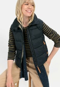 Schwarze Steppweste mit hohem Kragen, ausgestattet mit einem Reißverschluss vorne und Seitentaschen. Kombiniert mit einem gestreiften Langarmshirt und beigen Hosen.