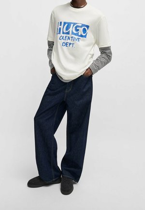 Personne portant un t-shirt blanc avec le texte bleu "HUGO CREATIVE DEPT." sur des manches longues rayées noir et blanc, un jean large et sombre, et des chaussures noires.