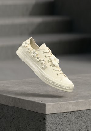 Baskets blanc cassé avec semelle en caoutchouc, décorées de petites appliques de fleurs 3D avec des centres en perles, flottant au-dessus d'un rebord en béton.