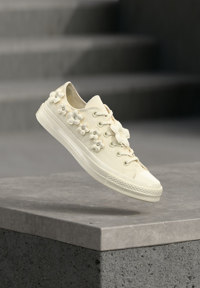 Ανθρακί αθλητικό παπούτσι με λαστιχένια σόλα, διακοσμημένο με μικρές εφαρμογές τρισδιάστατων λουλουδιών και κέντρα από χάντρες, αιωρούμενο πάνω από μια τσιμεντένια άκρη.