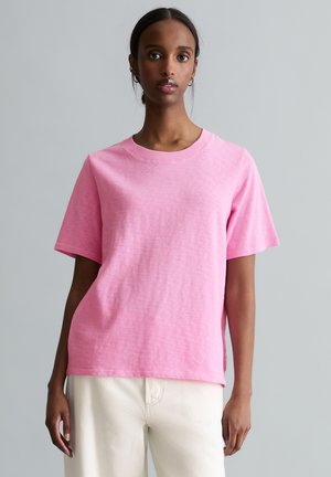 Kobieta ubrana w jednolity różowy t-shirt z krótkim rękawem i kremowe spodnie, stojąca na tle szarego tła.