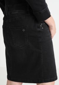 Personne portant une jupe en jean noire avec une poche arrière et de petits clous en métal en forme d'étoile près de la hanche, la main reposant sur le côté.