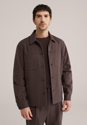 Lühikeste tumedate juustega mees, kes kannab pruunikat triibulist jopet ja sobivaid pükse, seisab tavalise tausta ees.