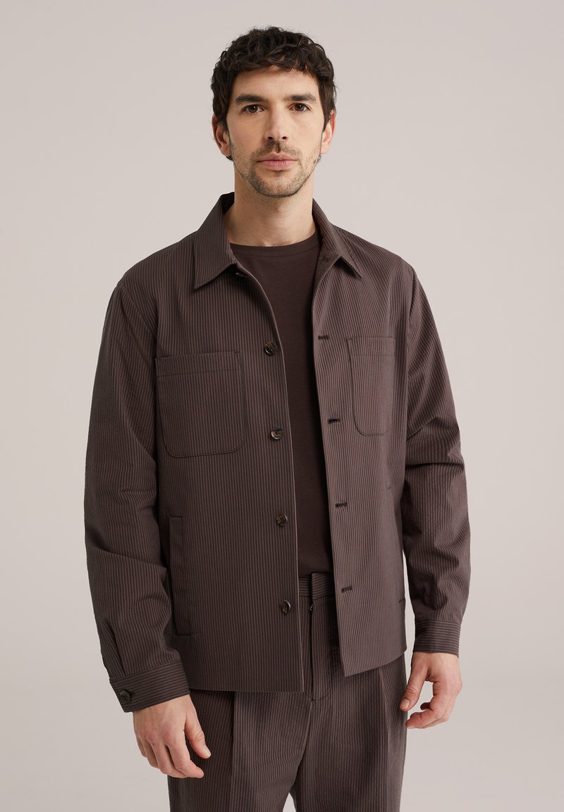 Mann mit kurzem dunklem Haar, der eine braune Nadelstreifenjacke und passende Hose trägt, vor einem einfarbigen Hintergrund stehend.