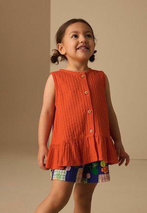 Uśmiechnięte małe dziecko z kitkami, ubrane w pomarańczowy bezrękawnik na guziki oraz kolorowe szorty w wzory, stojące na neutralnym tle.