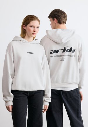 Deux jeunes adultes portant des sweat-shirts à capuche gris clair amples avec un texte noir ; l'un est face à l'avant, l'autre montre le dos du sweat-shirt avec un design graphique.