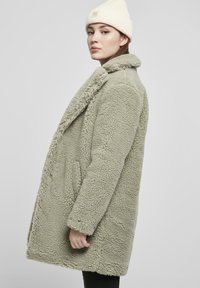 Svetlozelený, chlpatý fleece kabát s výstrihom do V, bočnými vreckami a voľným strihom, doplnený o krémovú pletenú čiapku.