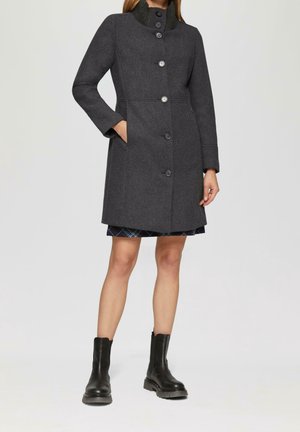 Kobieta ubrana w ciemnoszary płaszcz sięgający kolan z guzikami, spódnicę w kratę oraz czarne botki, stoi z jedną ręką w kieszeni.