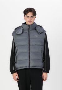 Grå polstret vest med høy krage, glidelås og to sidelommer. Har en glatt tekstur og lett design. Logo på brystet.
