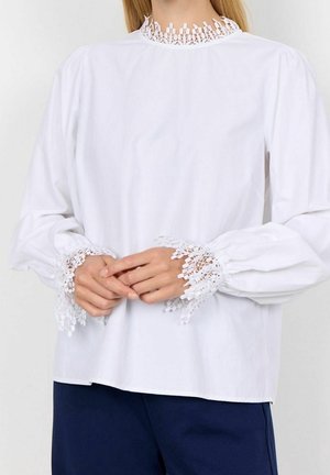 Woman wearing a white long-sleeve blouse with lace detailing on collar and cuffs, paired with dark blue pants. Hands clasped in front.