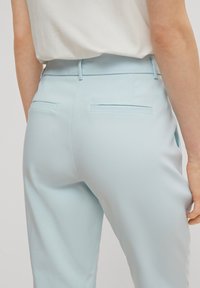 Pantalon bleu clair avec une texture lisse, doté de poches arrière et d'une coupe ajustée. La ceinture est plate avec des passants pour ceinture.