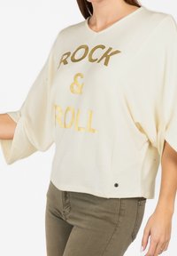 Maglietta in cotone bianco con lettering dorato "ROCK & ROLL". Presenta una vestibilità ampia, maniche larghe e un piccolo bottone decorativo sull'orlo.