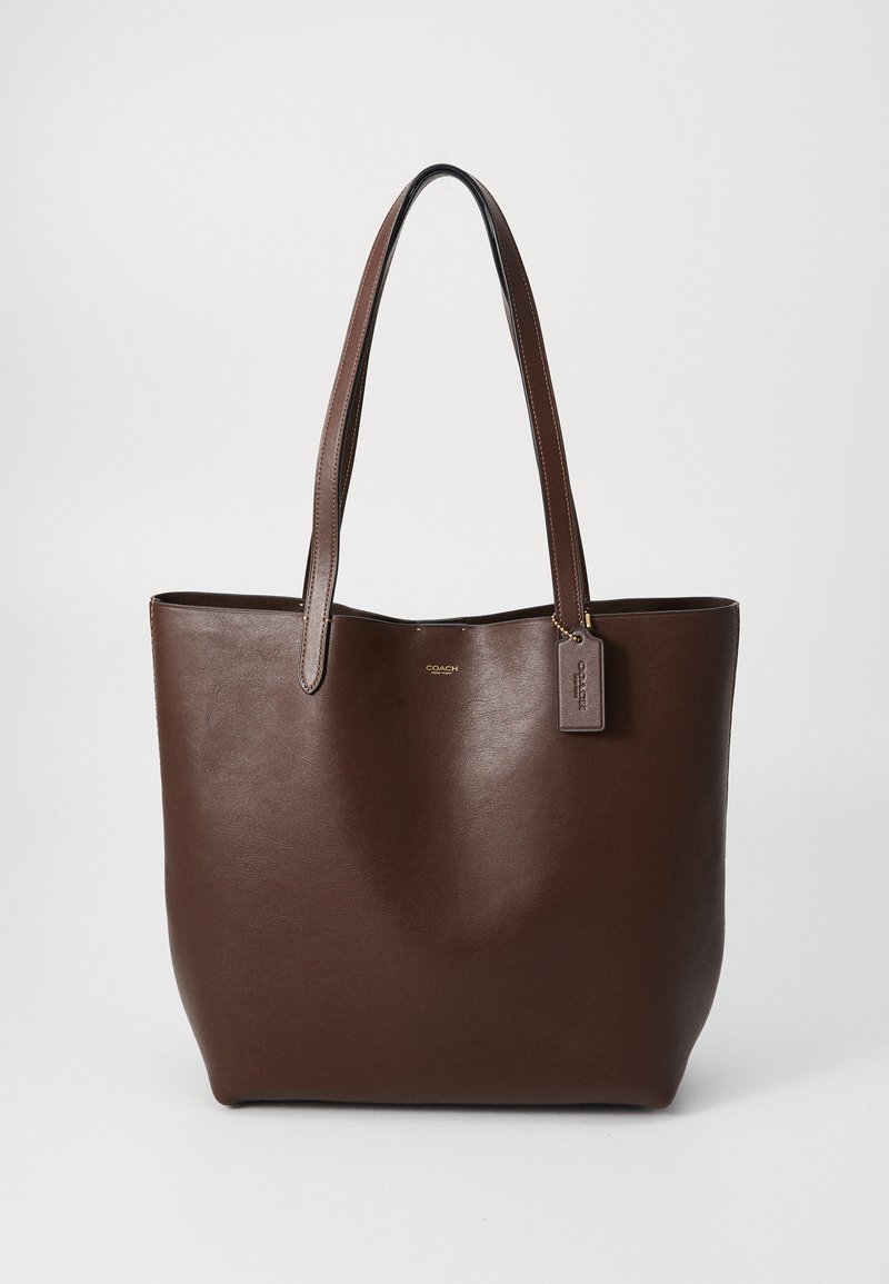 Borsa tote in pelle marrone scuro con due manici e un piccolo cartellino appeso su uno sfondo chiaro e uniforme.