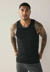 Black cotton tank top with a ribbed texture. Sleeveless design. Features a round neckline and form-fitting silhouette. Model stands in a neutral setting.