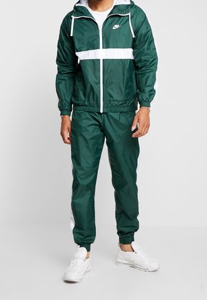 Mand iført mørkegrøn og hvid Nike træningsdragt med hættetrøje og hvide sneakers, stående på ensartet lyst baggrund.