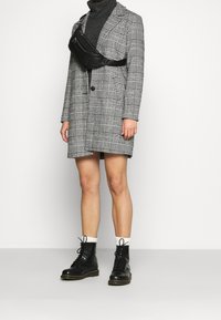 Cappotto a quadri grigi con un solo bottone, indossato sopra un dolcevita nero. Marsupio in ecopelle nera e stivali neri con lacci. Calzini bianchi.