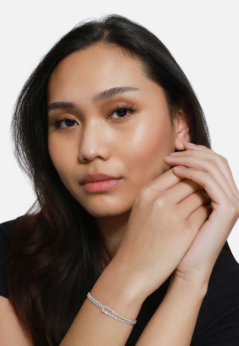 Bracciale d'argento intrecciato indossato su un polso, con una texture liscia. Il modello ha capelli scuri, pelle chiara e un trucco naturale.