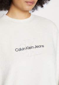Bílá mikina s výšivkou loga "Calvin Klein Jeans" v černé barvě, s jemnou texturou a kulatým výstřihem.