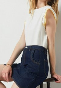 Bílý top bez rukávů s žlutým akcentem v švech, spárovaný s tmavými džíny sukní s volánkovou kapsou a oranžovým kontrastním prošíváním.