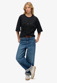 Camiseta negra de gran tamaño con un diseño gráfico texturizado, combinada con unos vaqueros holgados de color azul claro y zapatillas beige.