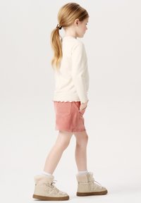 Lyscremefarvet skjorte med lange ærmer og bølget kant, parret med lyserøde kordshorts med lommer og beige støvler med pelsfor.