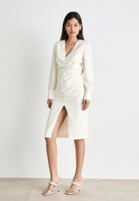 Cremiges Midikleid mit V-Ausschnitt, gerafftem Oberteil, langen Ärmeln, einem Schlitz vorne und gerippter Textur. Tragen mit nude-farbenen High Heels.