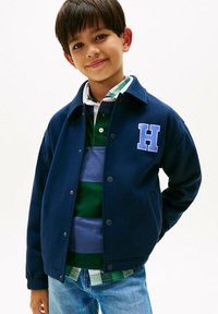 Ragazzo con una giacca blu navy con una toppa "H", maglietta polo a righe verdi e blu, e jeans, sorridente con una mano dietro la schiena.