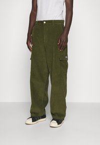 Person med olivgröna kordflöjelsbyxor och svarta och vita sneakers, som står mot en enkel vit bakgrund.