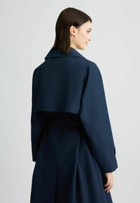 Cappotto trench blu navy con design cropped, ampio colletto e vita con cintura. Realizzato in tessuto liscio con una vestibilità su misura e particolare piega sul retro.