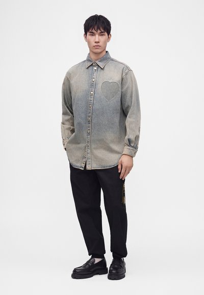Homme portant une chemise en jean grise délavée à boutons avec un détail cœur brodé, pantalon noir avec une fermeture éclair latérale et des mocassins noirs, debout sur fond blanc.