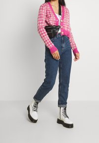 Růžovo-bílý kostkovaný zkrácený kardigán, doplněný o džíny s vysokým pasem v modré barvě a bílé šněrovací boty s černými podrážkami. Černý pouzdro na telefon nošené křížem přes tělo.