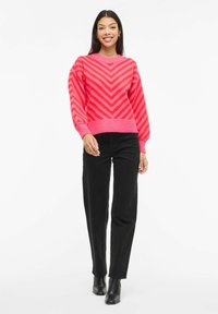 Pull rose avec rayures en chevron, poignets et ourlet côtelés, associé à un pantalon noir à jambes droites et des bottines. Texture lisse, coupe décontractée.