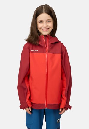 Junges Mädchen lächelt, trägt eine rot-bordeauxrote Mammut-Reißverschlussjacke und blaue Hose mit einem Logoaufnäher.