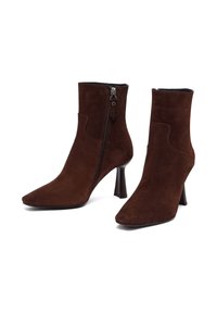 Stivaletti alla caviglia in suede marrone con punta a forma di freccia, zip laterale e tacco affusolato. Il materiale ha una trama morbida e un design elegante.