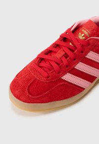 Sarkanas zamšādas sneaker ar rozā ādas svītrām, teksturētu mēliti un zelta logotipa akcentiem. Aprīkots ar apaļām auklām un gumijas zoles gumijas zoli.