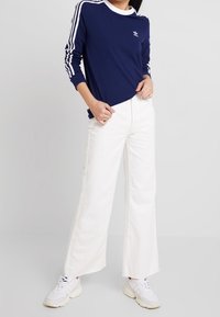 Laivastonsininen pitkähihaisten paita, jossa valkoinen kaulus ja kolme valkoista raitaa hihoissa, yhdistettynä valkoisiin leveälahkeisiin housuihin ja valkoisiin tennareihin.