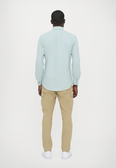 Chemise à manches longues bleu clair avec des poignets à boutons, associée à un pantalon cargo beige et des baskets blanches, vue de dos.