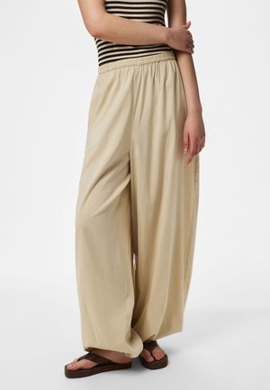 Osoba nosząca beżowe spodnie o szerokich nogawkach, koszulkę bez rękawów w paski oraz brązowe sandały, stoi z jedną ręką skrzyżowaną na ciele.