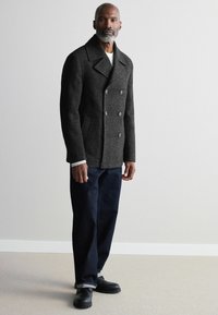 Abrigo gris tipo peacoat con botones de doble pecho, grandes solapas y dos bolsillos laterales, llevado sobre una camisa de rayas y jeans oscuros con dobladillos.