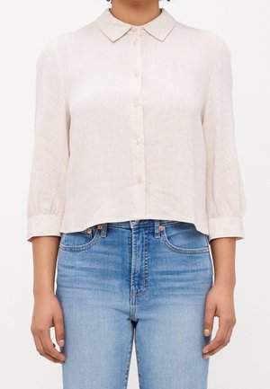 Femme portant un chemisier beige clair à col boutonné avec manches trois-quarts et un jean en denim bleu taille haute sur fond blanc.