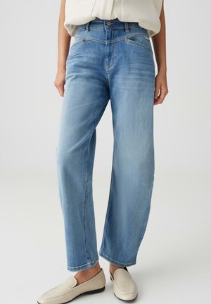 Frau trägt hellblaue High Waist Jeans mit weitem Bein, weiße Bluse in die Hose gesteckt und beigen Slipper, steht vor einem einfarbigen Hintergrund.
