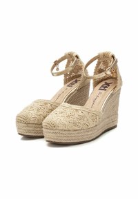 Sandalias de cuña beige con parte superior de tejido trenzado, intrincados detalles de cuerda y correa al tobillo con hebilla de metal. Suela de yute texturizada.