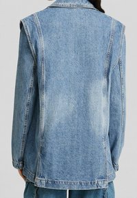 Denimová vesta v světle modré barvě, s ramenními švy, viditelným prošíváním, uvolněným střihem a mírně opotřebovanou texturou na zadní části.